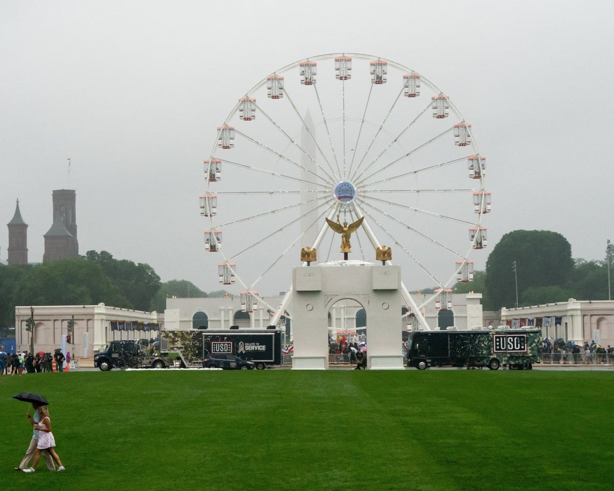 People under an umbrella cross a lawn in front of a Ferris wheel