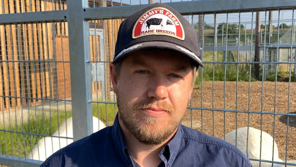 A man standing in front of a fence with an animal enclosure beyond it. He is wearing a blue shirt and a baseball cap, with a 