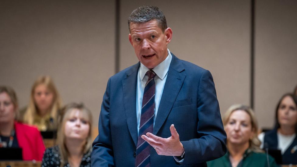 Rhun ap Iorwerth is stood in the Senedd with a group of Plaid MSs behind him, wearing a suit.