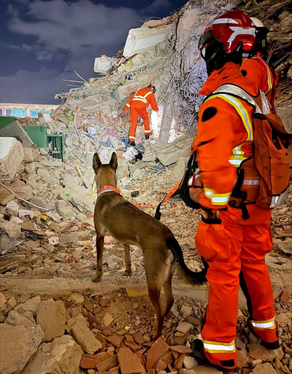 A person dressed in head-tot-toe orange hi-vis holds a dog on a lead, as they look for survivors in an area covered in rubble in La Guaira, Venezuela.