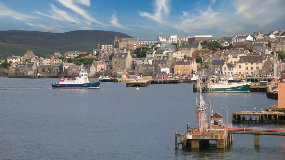 Wide view of a coastal harbour with several boats on calm water, including a ferry and fishing vessels, alongside a town of stone buildings on a gentle hillside under a blue sky with scattered clouds.