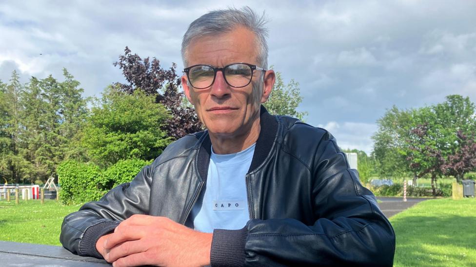 A man is sat at a park bench with his hands resting on the table, clasped in front of him. He has short dark hair which is turning largely grey. The man, dressed in a blue tshirt and black leather jacket, is looking at the camera through his large, round-rimmed glasses. Behind him is a footpath surrounded by grass on both sides, some trees and a children's play park in the distance. 