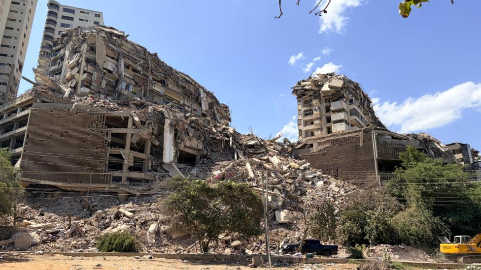 Dramatically collapsed buildings behind a dusty roadside lined with trees