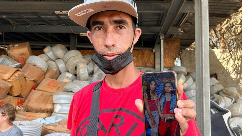 A man in a baseball cap and red t-shirt displays a photo of his wife and daughter on a mobile phone he holds aloft in his left hand.