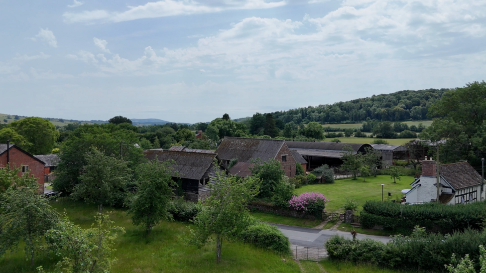 The farm includes historic timber barns, and modern buildings. There is a black and white cottage and an orchard next to the old barns.
