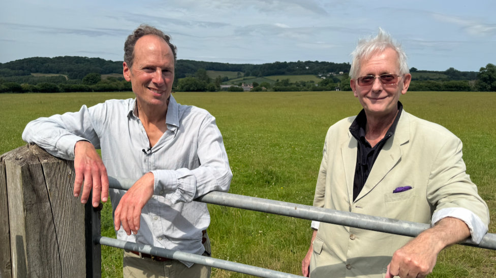 The two men are leaning on a gate. In the background are the farm's meadows, with hills beyond. 
