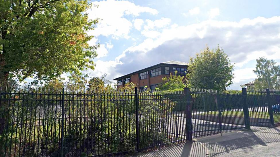 A school building set against bright skies is behind high black metal fencing. There are bushes and trees in the grounds of the school.