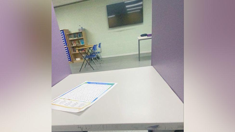 A low-quality photo, taken slightly askew, from the viewpoint of someone sitting at a desk with two lilac-coloured plastic dividers blocking the view to the sides. Straight ahead can be seen a view of what appears to be a sparsely furnished classroom, with a couple of desks, a small bookshelf and a TV on the wall. On the desk in the booth is a word-search.