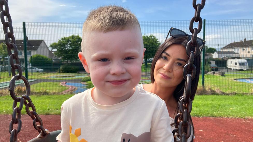 Gemma McConnell, a woman with long, straight brown hair, pushing her four-year-old son Dáibhin on a swing in a playpark on a sunny day. She is standing behind her son, wearing sun glasses on her head. He is smiling and wearing a beige t-shirt adorned by yellow and brown dinosaur motifs. There are houses and vehicles in the background. 