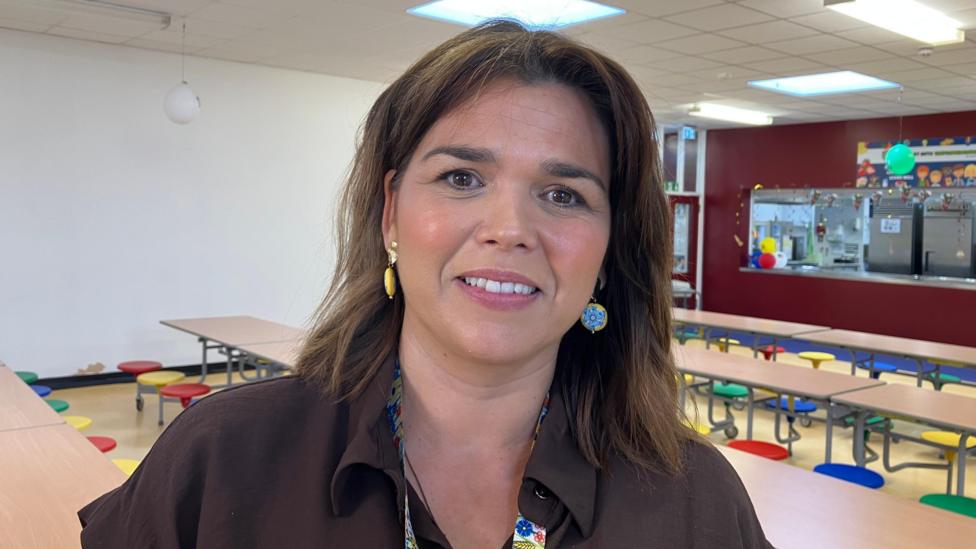 Catherine Dourish, a woman with shoulder-length brown hair, stands in a classroom smiling at the camera. She is wearing a brown blouse and colourful earrings. There are wooden desks with multi-coloured seats in the background. 
