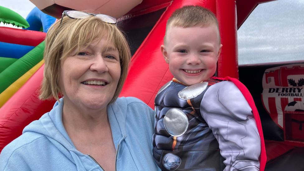 Dáibhin McConnell-Mailey, a four-year-old boy with short, blond hair, being held up by his nursery teacher Mary McLaughlin. She has a short blonde bobbed hairstyle and is wearing reading glasses on her head and a pale blue hooded top. Dáibhin is wearing a grey superhero costume with a red cape. They are pictured in front of an inflatable slide. 