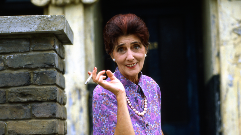June Brown as Dot cotton in Eastenders. She is wearing a purple shirt, has short permed hair and is holding a cigarette