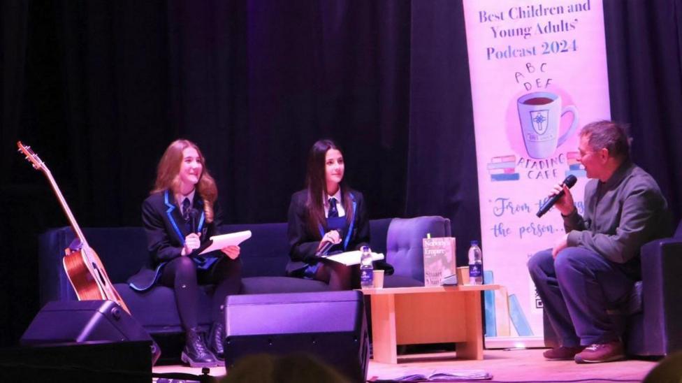 Two female school pupils sit on a couch carrying out an interview with Stuart Murdoch - a man with short hair, holding a microphone. 