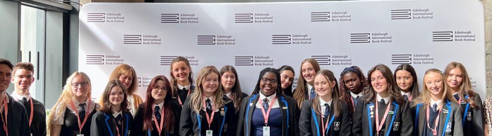 A group of smiling school pupils, standing in front of banners and signs advertising the Edinburgh International Book Festival.