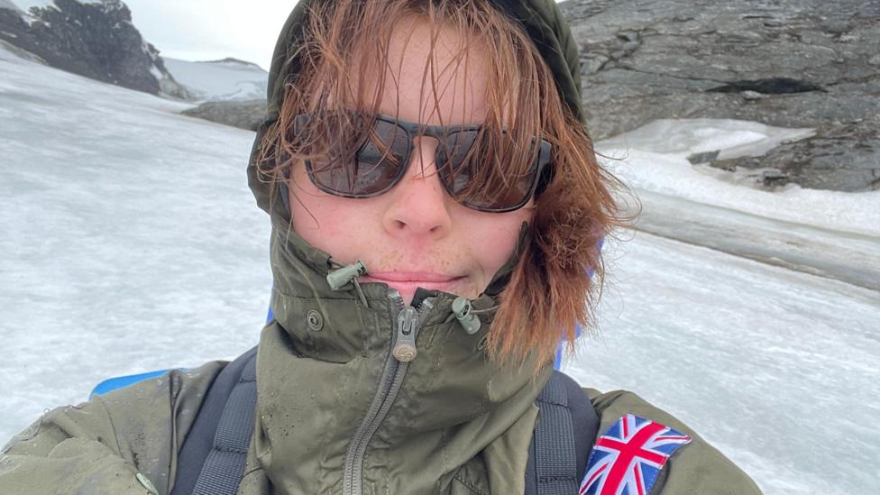 A selfie of Emily Broadwell wearing a khaki raincoat and black sunglasses with her hood up, and her gingery-brown hair poking out. Snow and ice on rocky mountains surround her.