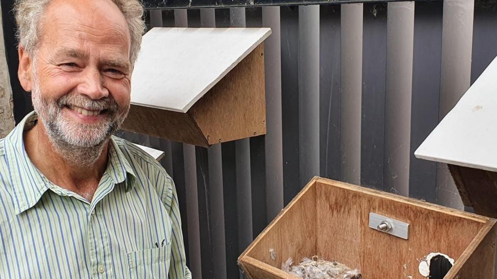 Dr Mike Rands is smiling at the camera standing in front of two wooden bird nesting boxes. He has greying hair and facial hair and is wearing a striped shirt.