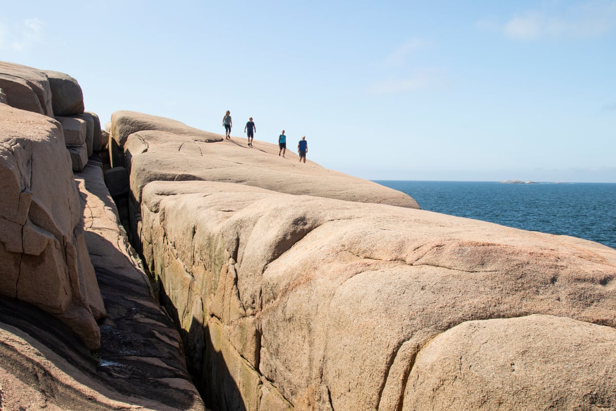 A huge granite cliff by the sea with four walkers in the distance