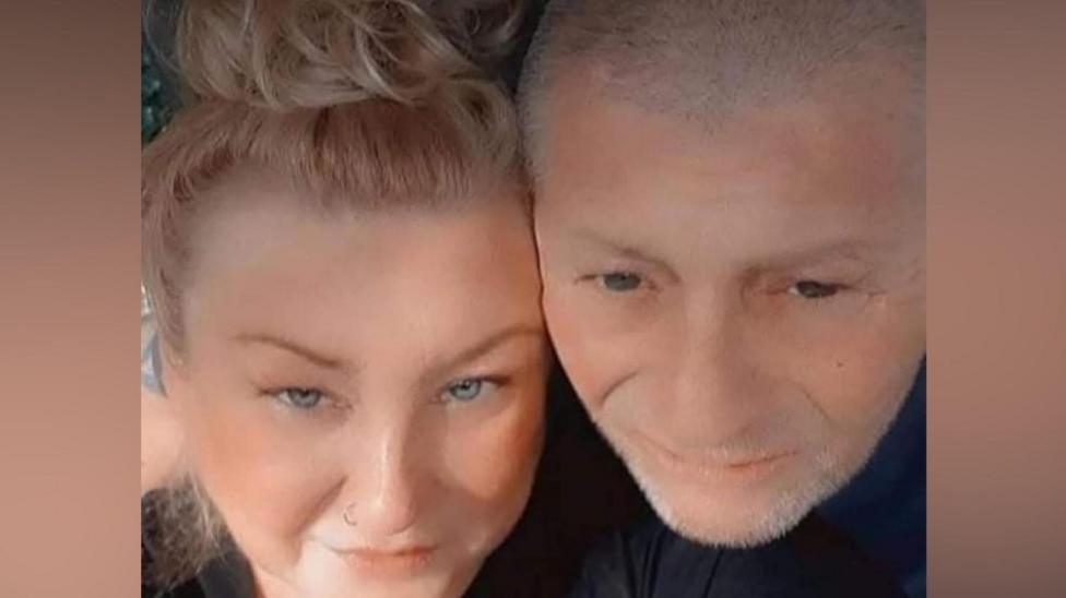 A selfie showing Suzanne Hall, who has blonde hair pulled back into a ponytail, cheek to cheek with her husband, who had short grey hair and grey stubble