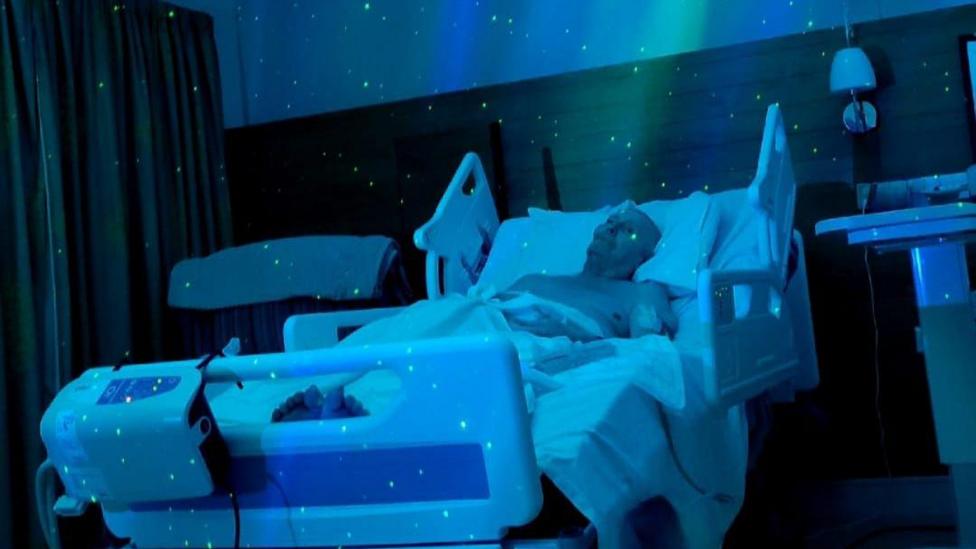 A shirtless man lies in a hospital bed, propped up on a pillow, while blue and green sensory lighting surrounds him