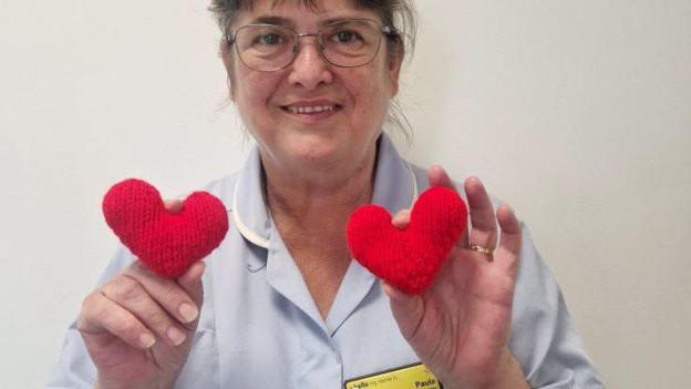 A woman with brown hair cut into a fringe and glasses, and wearing a light blue hospice uniform with a name badge reading 'Hello my name is Paula' holds up two small knitted red love-hearts to the camera, one in each hand. 