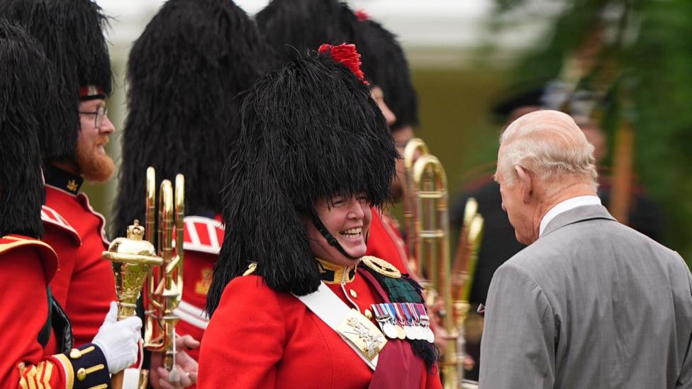 King Charles III (right) during the Ceremony of the Keys in the gardens of the Palace of Holyroodhouse in Edinburgh. He has his back to the camera. Facing the camera are members of the band of the Royal Regiment of Scotland. They are wearing red and gold jackets, a black feather bonnet and are carrying gold instruments.