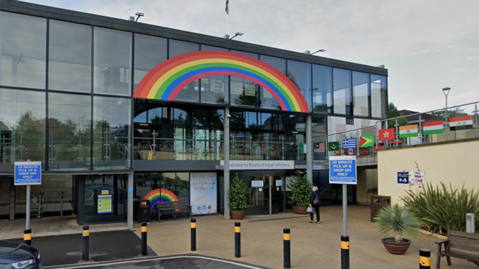 A three-floor glass hospital building with a rainbow painted on the outer windows of the top floor. There are flags on the walkway to the right