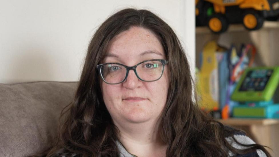 A woman with round glasses and long brown hair sitting on a sofa.