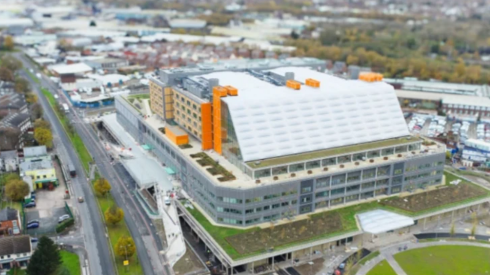 Aerial view of a very large rectangular building, with grey floors at the bottom and a white roof with orange elements to the side and top.