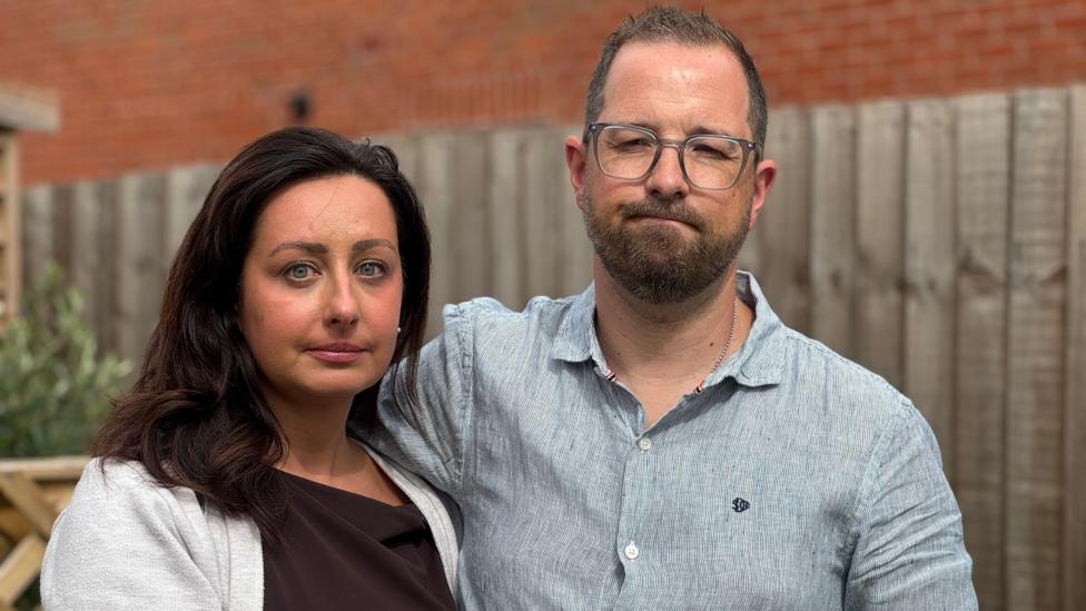 A woman and a man stand in front of a wooden fence, pictured from the shoulders up. The woman has shoulder-length dark hair and is wearing a brown top and grey cardigan. The man, who has a beard and brown hair, is wearing a blue shirt and glasses.