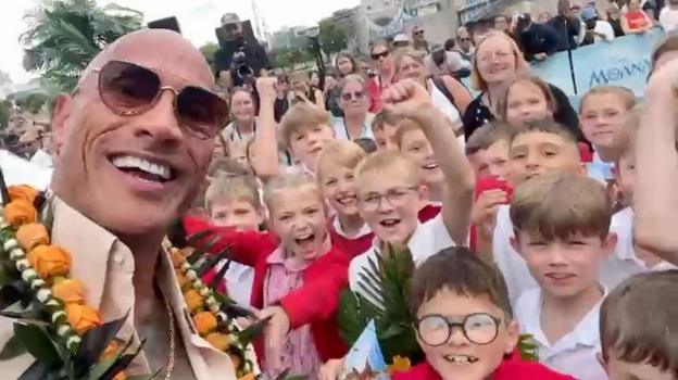 A selfie-style image taken by The Rock – a man wearing glasses and smiling. Next to and behind him is a group of schoolchildren. They are cheering, smiling and shouting. They are wearing red and white school uniforms.