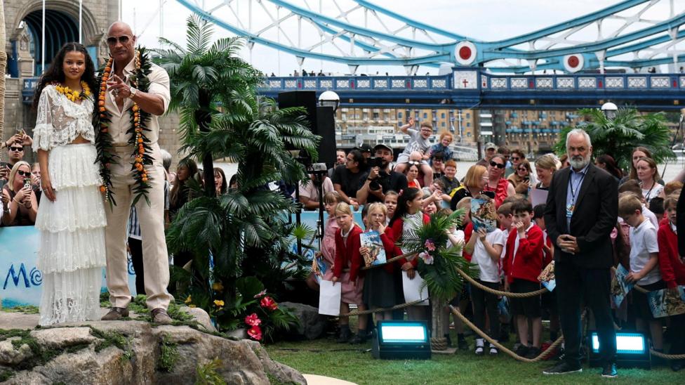 A woman and a man standing on top of a rock in front of a tree. To the right of the picture is a group of schoolchildren wearing red uniforms. They are standing behind a rope. In the distance is a bridge.