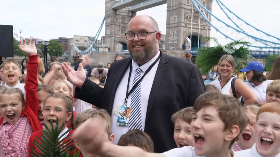A man wearing a black suit blazer over a white shirt with a striped tie stood in the middle of a group of schoolchildren who are wearing white and red uniforms. In the background is Tower Bridge.