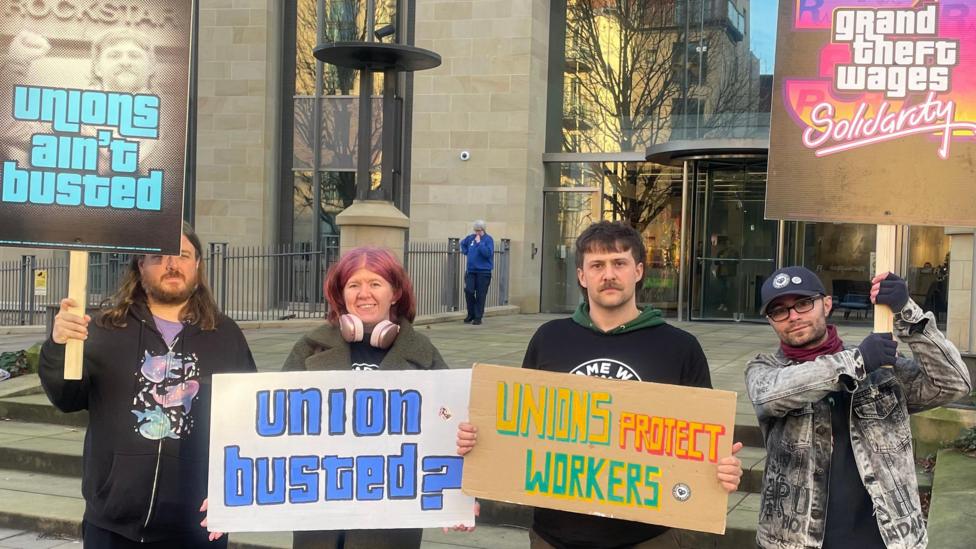 Four people, three men and a woman, hold signs outside a building. Their signs read 