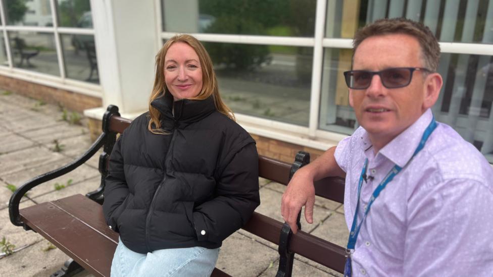 Lowri and Dr Healy sitting on a bench. Dr Healy is wearing a purple shirt with a lanyard, he has short dark hair and wears glasses. Lowri is wearing a black puffer jacket and jeans. 
