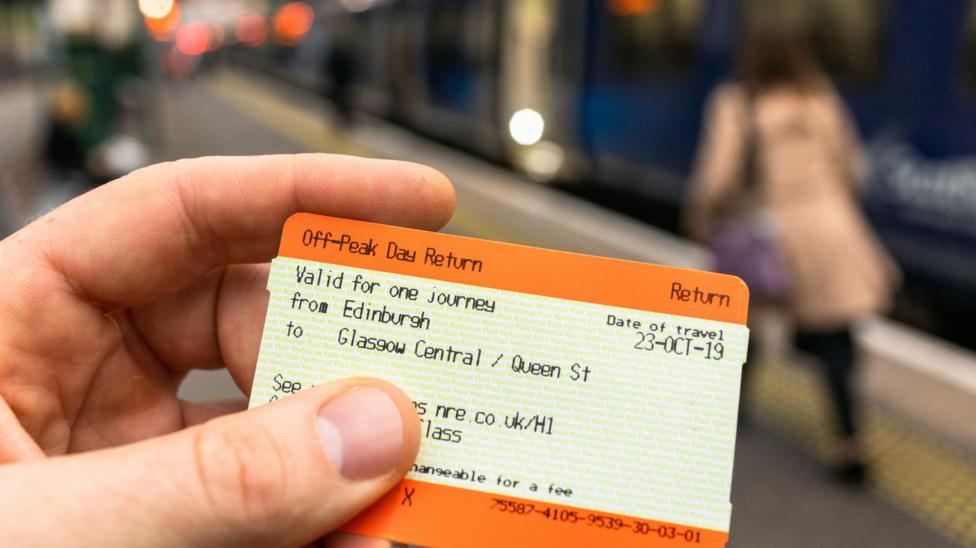 A hand holding a train ticket. There are people on the platform in the background which is blurred.