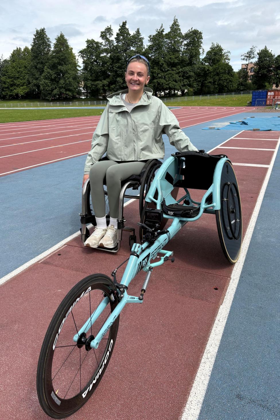 Portrait image of Melanie Woods with her racing wheelchair. She is on running track. She has brown hair tied back and black sunglasses on the top of her head. She's wearing a light khaki green rain jacket and a grey t-shirt and grey leggings. Her wheel chair is has a pale blue frame with two black wheels at back and one at the front