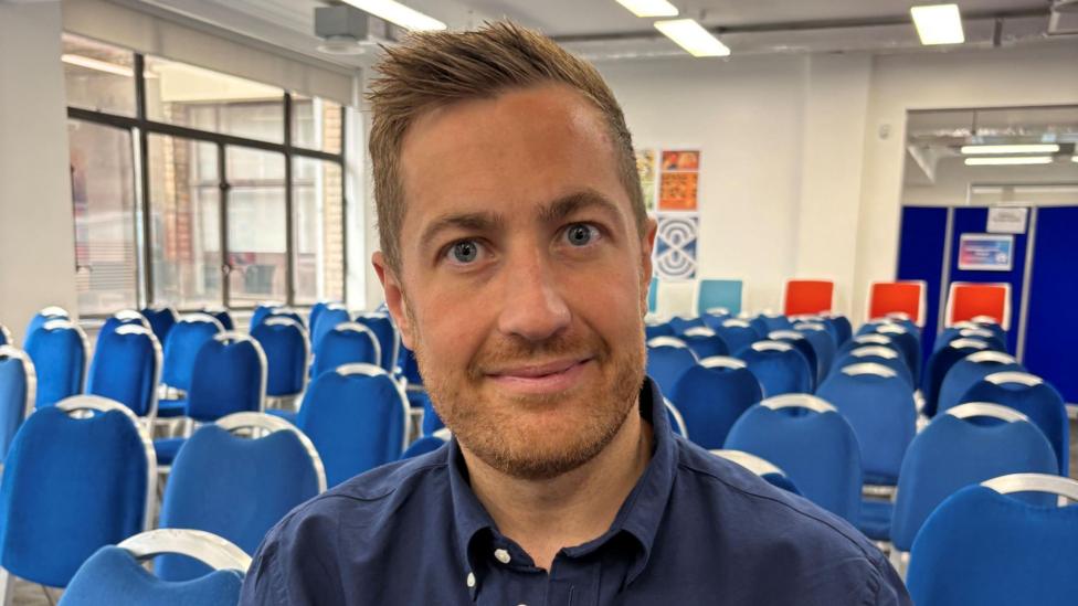 Head and shoulders image of Glasgow 2026 chief Phil Batty. He's sitting in a room full of blue chairs. He's looking directly at the camera and has spikey strawberry blond hair with a reddish goatie beard and moustache. He is wearing a navy blue shirt.