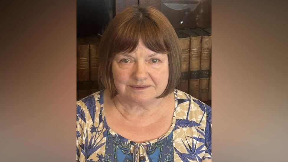 Colette Breen. She has shoulder-length brown hair with a fringe and brown eyes, wearing a yellow and blue patterned blouse.