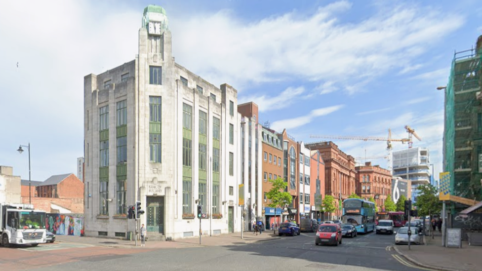 Image of the former bank of Ireland in Belfast. It is a tall white art deco building with green detailing standing at a street corner in front of a junction. Traffic, including buses and cars, moves along the road beside rows of red‑brick and modern buildings under a bright, partly cloudy sky.