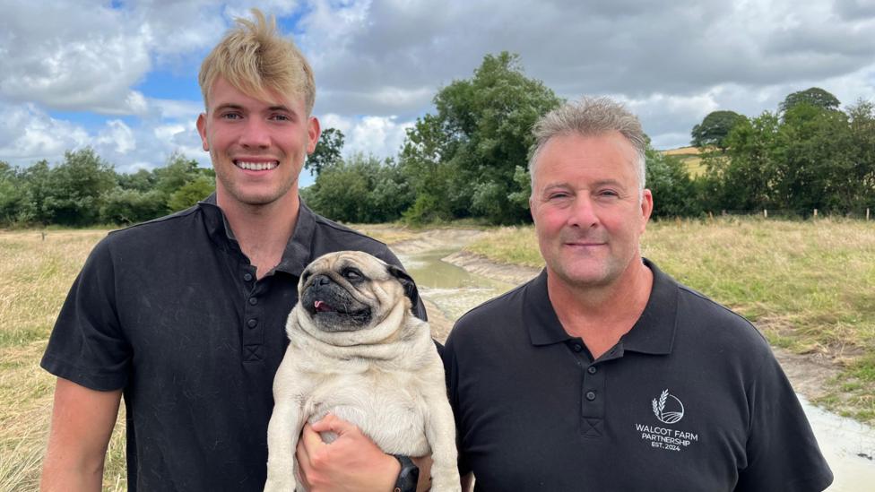 Will and Rob stood side by side, both wearing black polo shirts. Will has short blond hair, and Rob has short grey hair. Will is holding a black-and-tan pug dog. They are both stood on the bank of the new meander and are smiling widely.