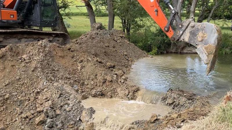 A large orange digger perched on an earth bank, with the scoop pointing downwards towards the water. Water rushes through where the digger has lifted out soil, with the water starting to pool in places as it flows into the new channel. The banks are lined with trees and shrubbery.