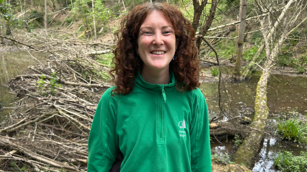 A woman with red curly hair is standing in front of a number of trees in a forest. She is wearing a green jacket with a tree logo and