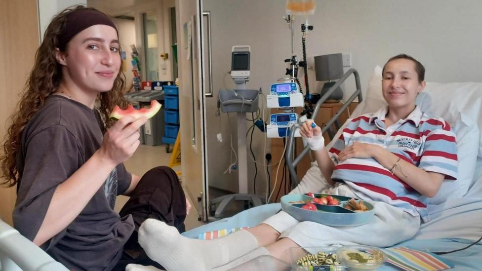 Grace and Caitlin in hospital. Grace has long dark hair and is wearing a dark top. Caitlin has short hair and is wearing a stripey top