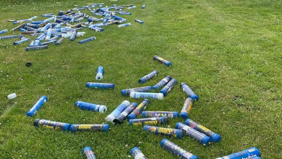 Blue Cream Deluxe canisters are lying all over a field. They are not in one pile but spread out.