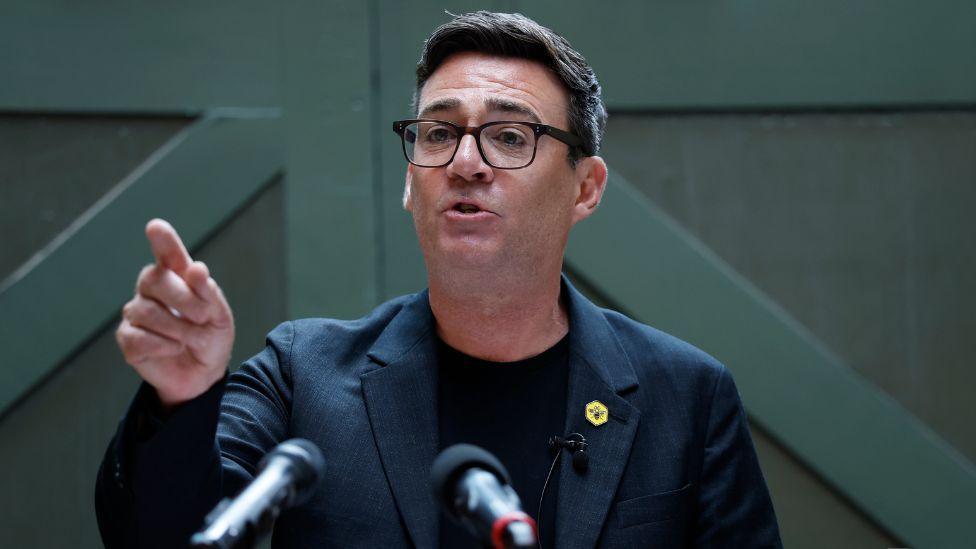 Andy Burnham, who has dark hair, speaks with his right hand raised and his index finger pointing ahead. He is wearing a dark suit and t-shirt 