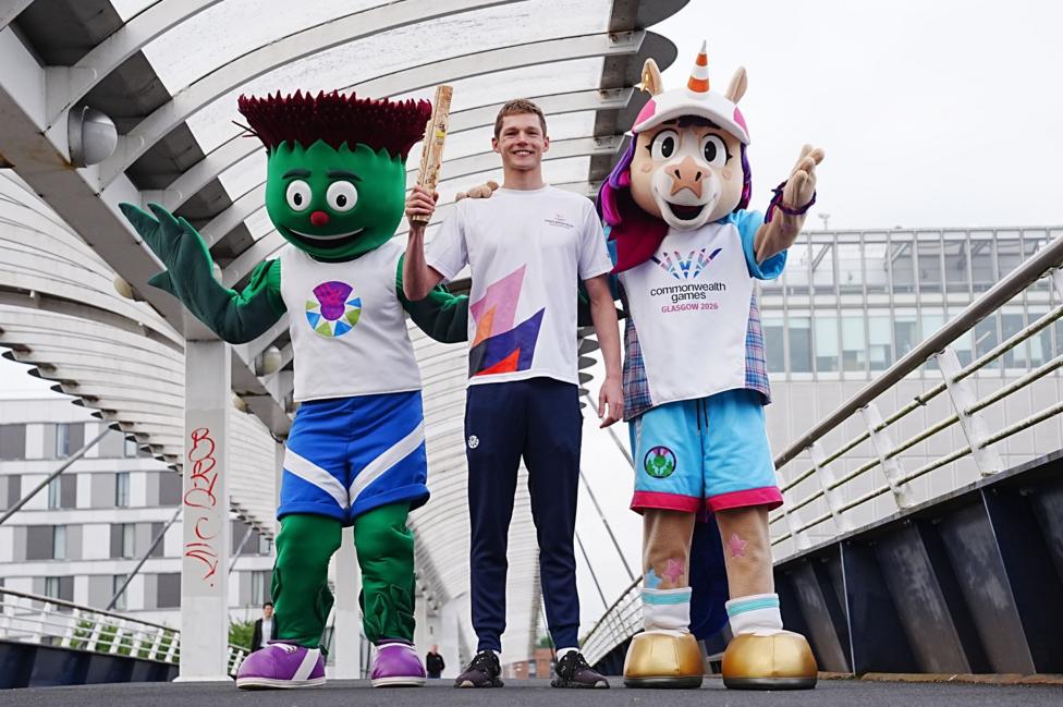 Two mascots standing either side of a young man holding a wooden baton on a bridge. The mascots are a thistle and a unicorn, both with smiling faces and wearing Team Scotland uniforms.