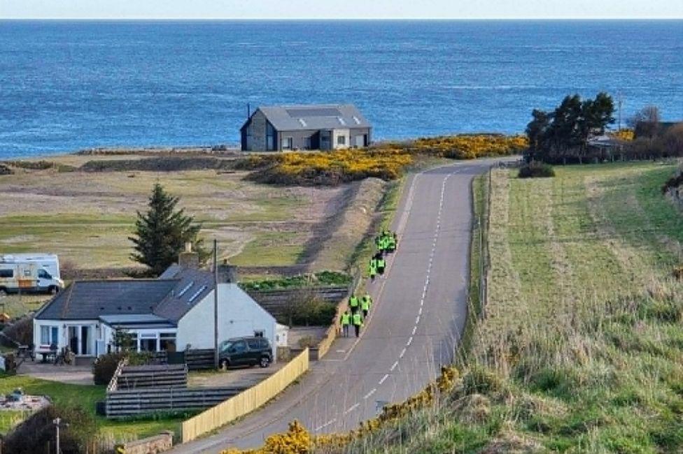 Children in hi-vis jackets walking on a rural footpath next to a road, with fields on either side, and the North Sea in the distance.