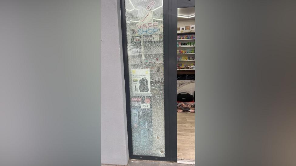 The broken window of the shop. The window is in a grey frame and the glass is shattered.