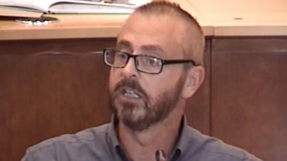 A blurry image of a man with short dark hair and a beard, wearing glasses and a grey shirt.