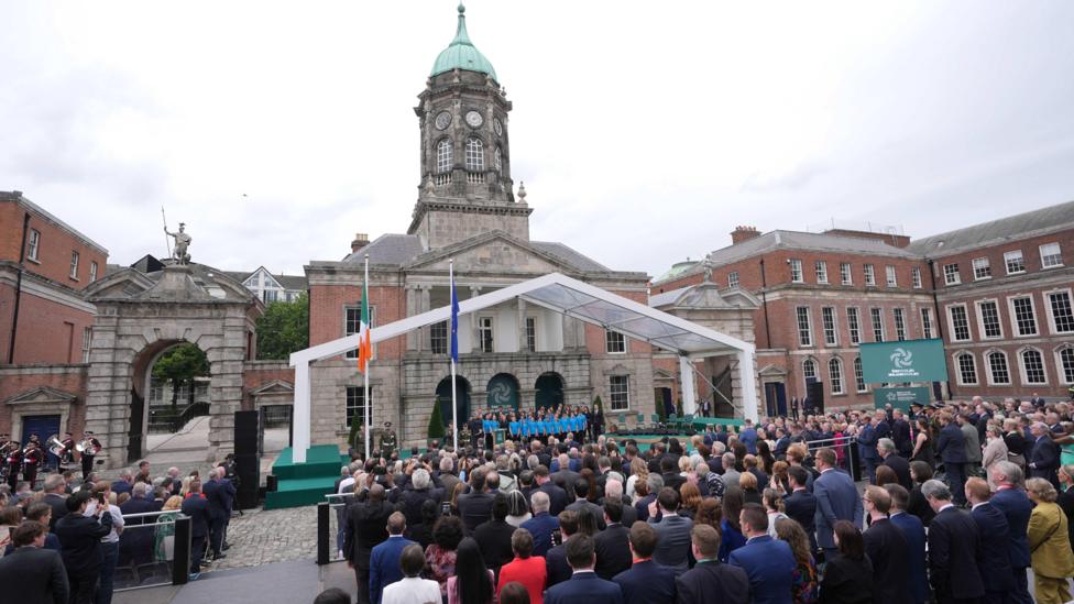 A large crowd seated in an urban area. In front of the crowd there is an Irish flag and an EU flag. Many of the crowd are wearing suits. The sky is overcast, and the area is surrounded by red brick buildings.